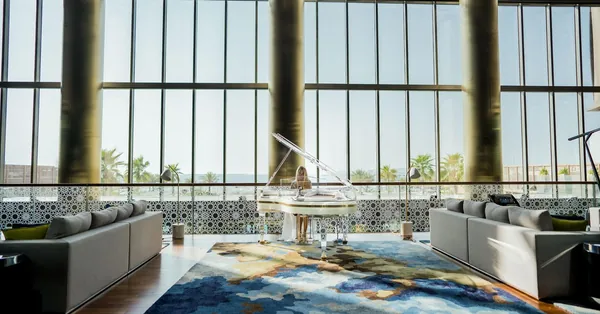 A hotel lobby with a glass wall, grey sofas and a white piano on a blue rug
