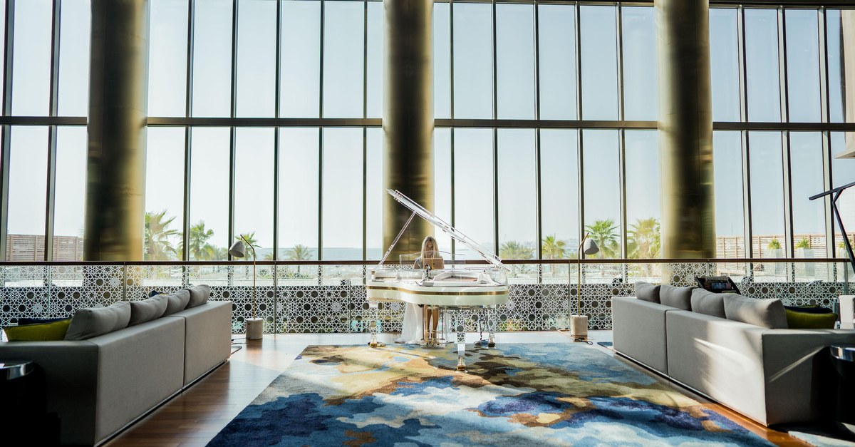 A hotel lobby with a glass wall, grey sofas and a white piano on a blue rug